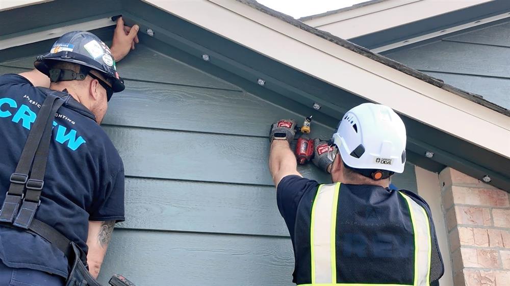 Two installers in hard hats and safety vests fastening mounting channel under the eave of a gray-sided home